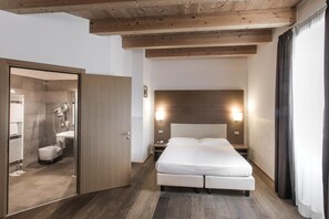 A hotel room with a wooden ceiling, a large bed with white bedding, a nightstand with a lamp, and a bathroom visible through an open door.