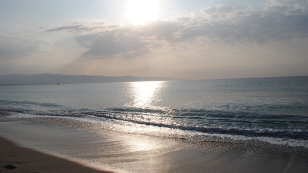A beach with sunlight reflecting off the water, a clear sky, and distant mountains. - Lewisrooms Domo (Cagliari)