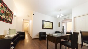 A spacious living area with a wooden floor, a dining table with chairs, a black sofa, and a decorative wall hanging. - Lewisrooms Domo (Cagliari)