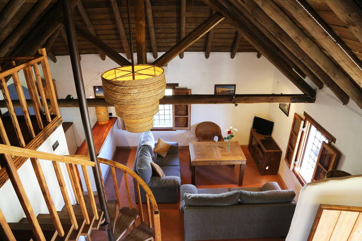 A loft-style room with a wooden staircase, a sofa, a dining table, and a hanging basket.