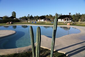Une piscine bordée de sable, entourée de cactus et de chaises longues bleues.