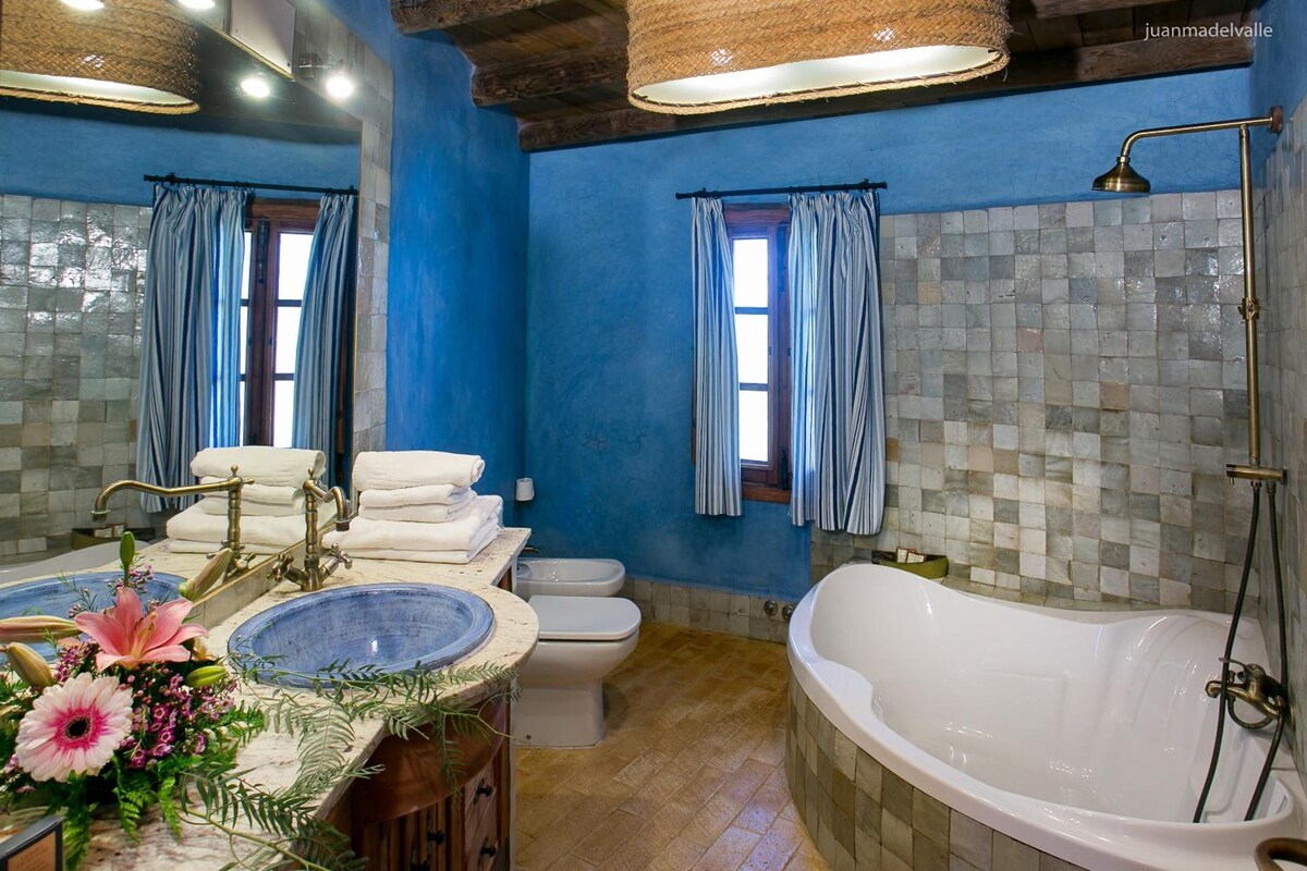 Un baño con bañera, lavamanos e inodoro. Hay cortinas en las ventanas y las paredes están pintadas de azul.