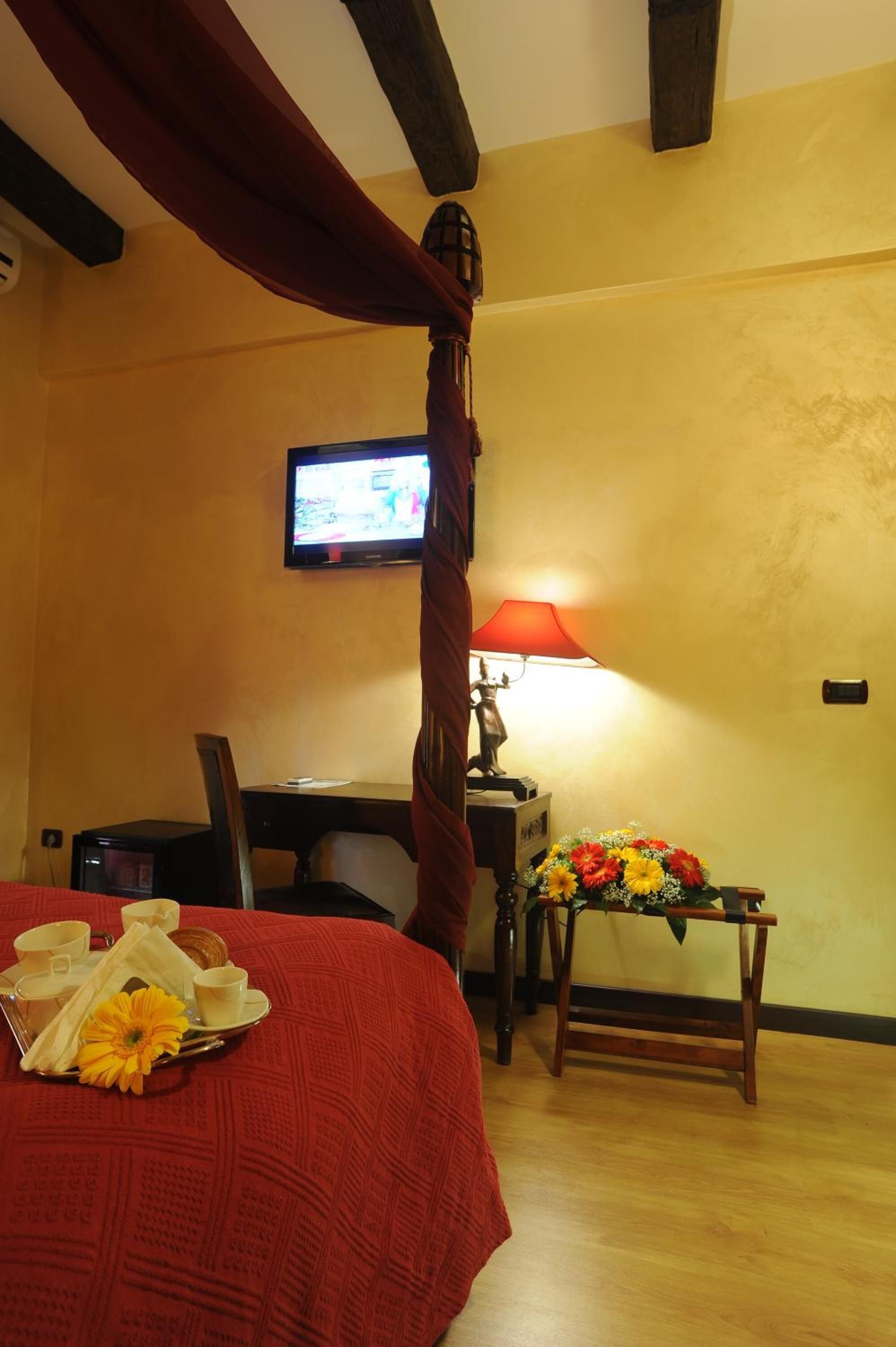 Una habitación de hotel con cama, escritorio, televisión, lámpara y una pequeña mesa con flores.
