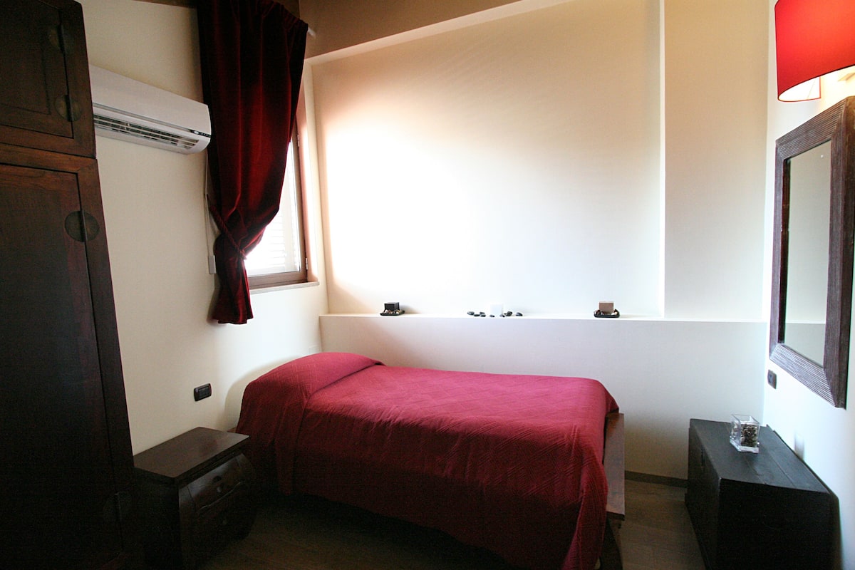 Een kleine, eenvoudige kamer met een bed, nachtkastje, spiegel en een raam met gordijnen.