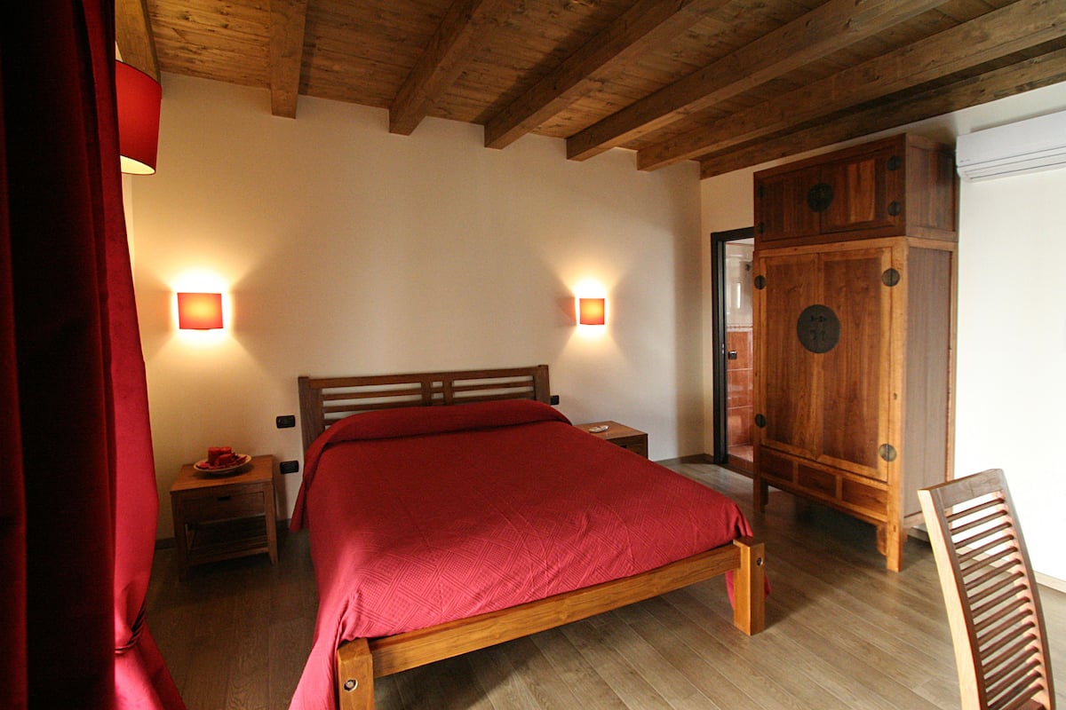 Een houten bed met een rode spreidek en een kamer met houten balken in het plafond en twee wandlampen.