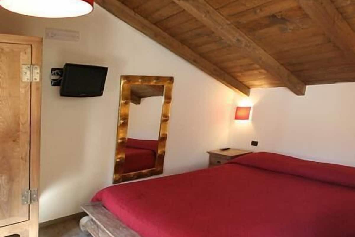 Een slaapkamer met een houten plafond, een bed met een rode sprei, een aan de muur bevestigde televisie en een houten kast.
