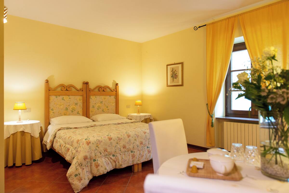 Um quarto com uma cama, mesas de cabeceira, uma cadeira, uma janela com cortinas e um vaso com flores.