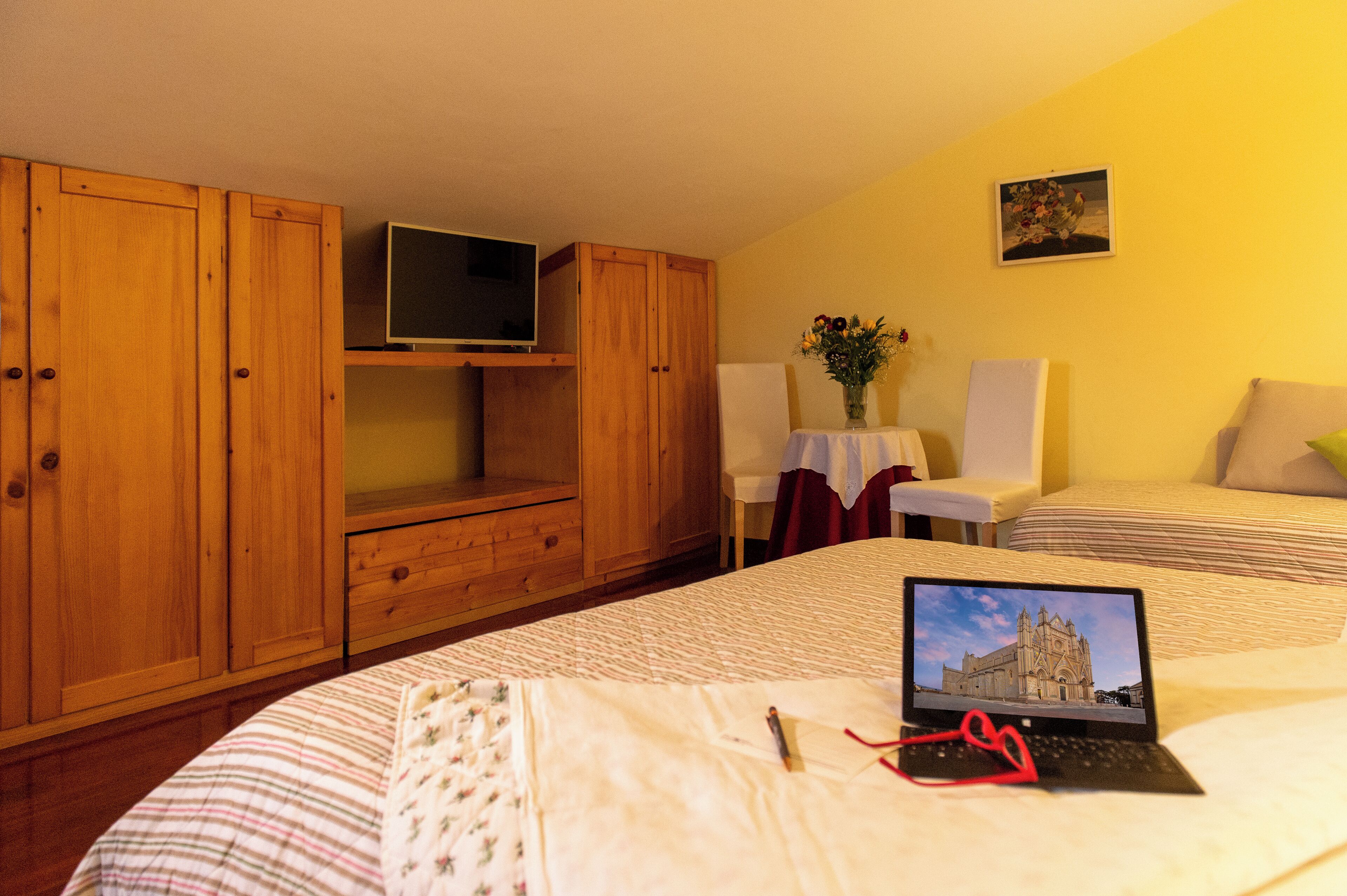Una camera da letto con un letto, un laptop, una TV su uno scaffale, una sedia e un tavolo con un vaso.