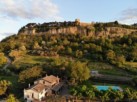 A villa with a swimming pool, surrounded by lush greenery and a cliff with a tower.