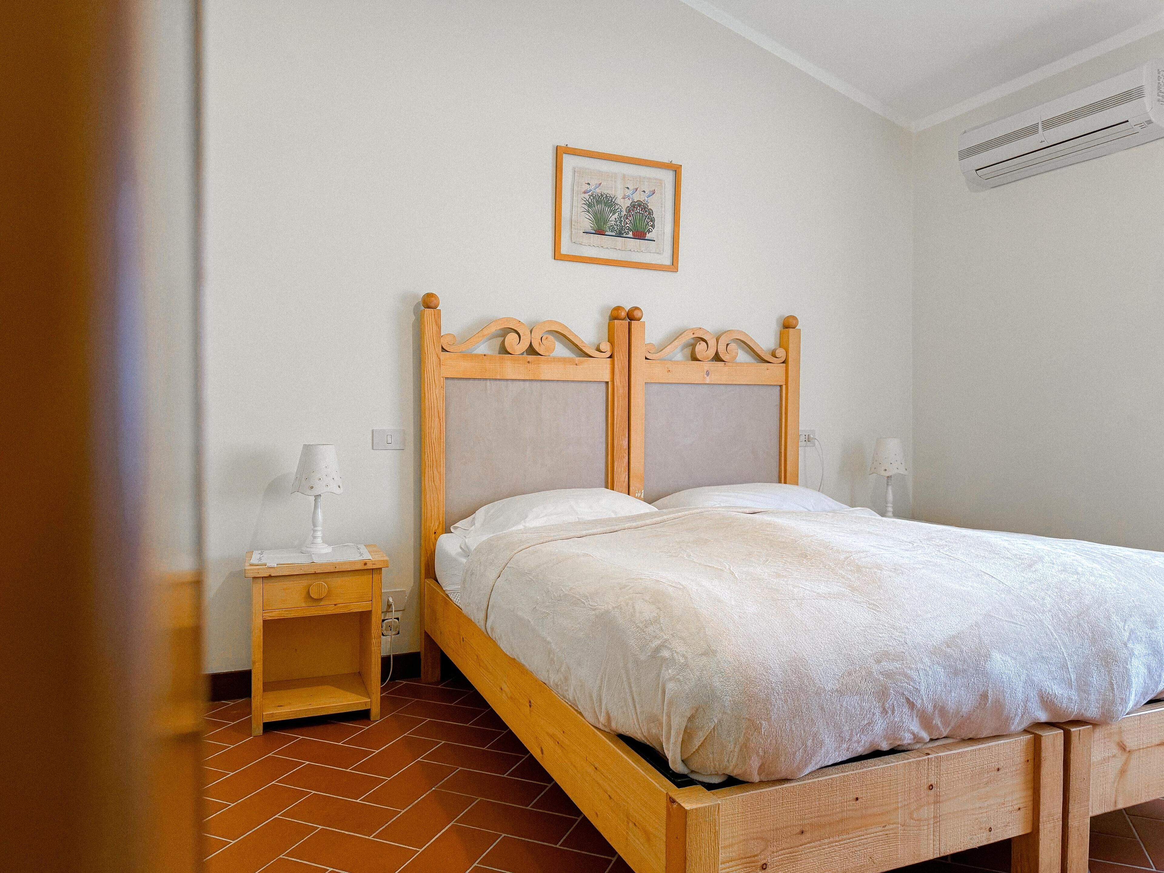 A wooden bed with a headboard, a nightstand with a lamp, a framed picture on the wall, and an air conditioning unit.