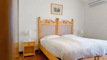 A wooden bed with a headboard, a nightstand with a lamp, a framed picture on the wall, and an air conditioning unit.