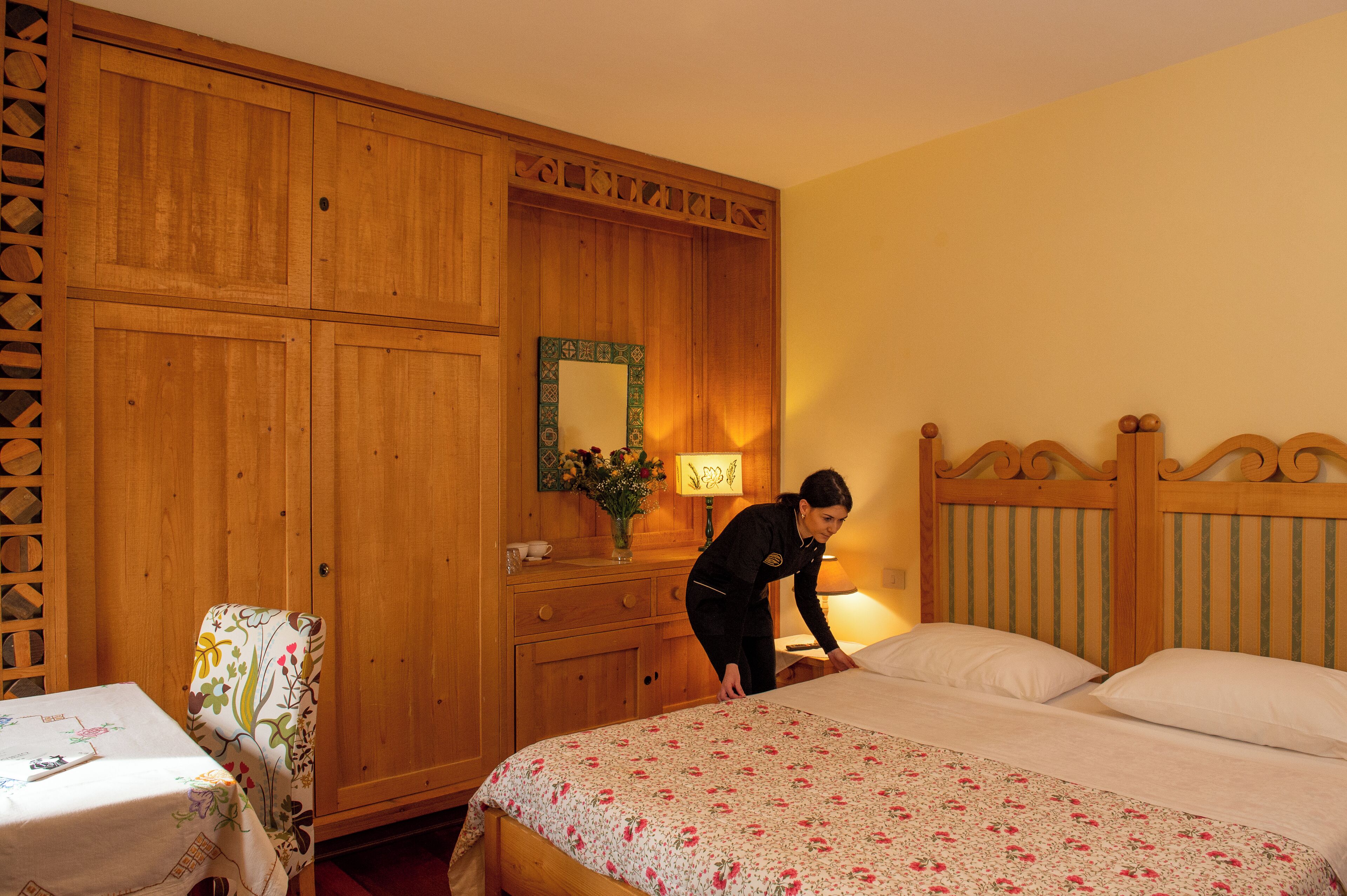 Habitación de hotel con un armario de madera, una cama con ropa de cama floral, una silla con funda floral y una mesita de noche con lámpara.