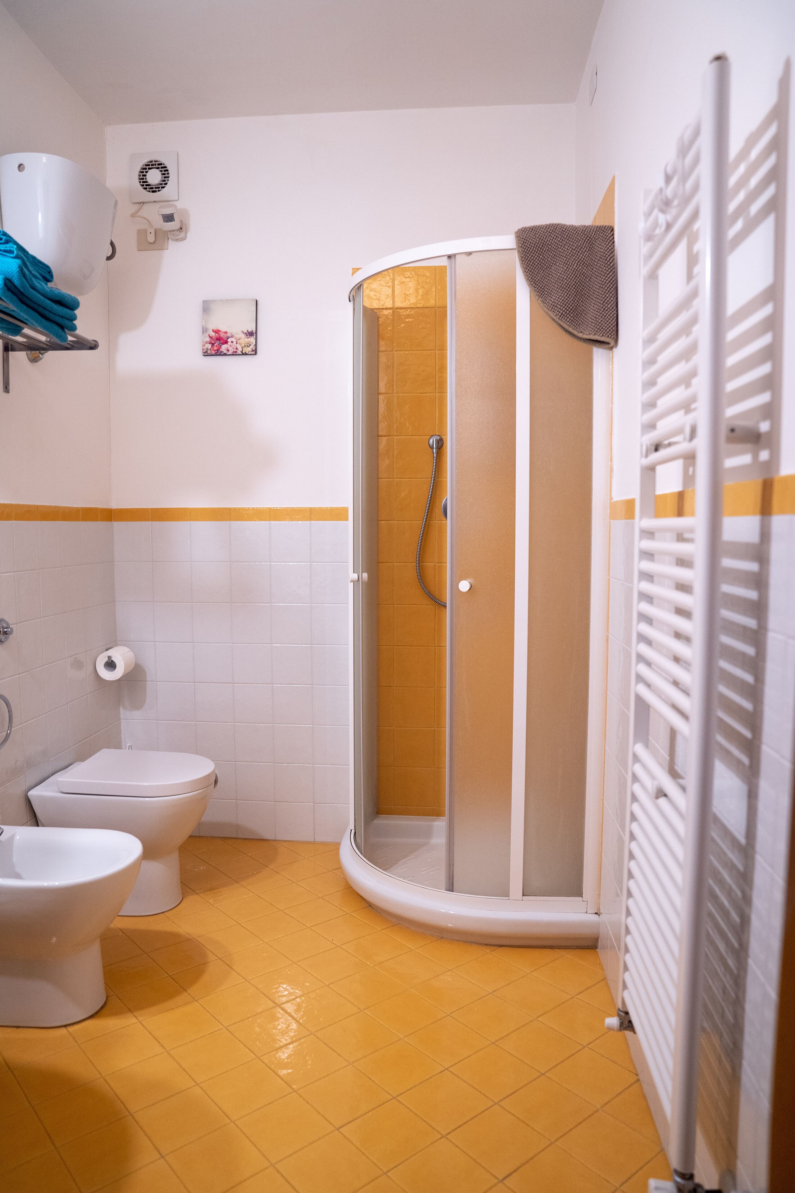 A bathroom with a shower, two toilets, a towel rack, and a radiator.