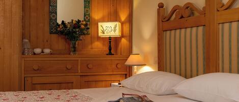 A wooden bed with white bedding, a bedside lamp, a mirror, and a dresser with a vase of flowers.