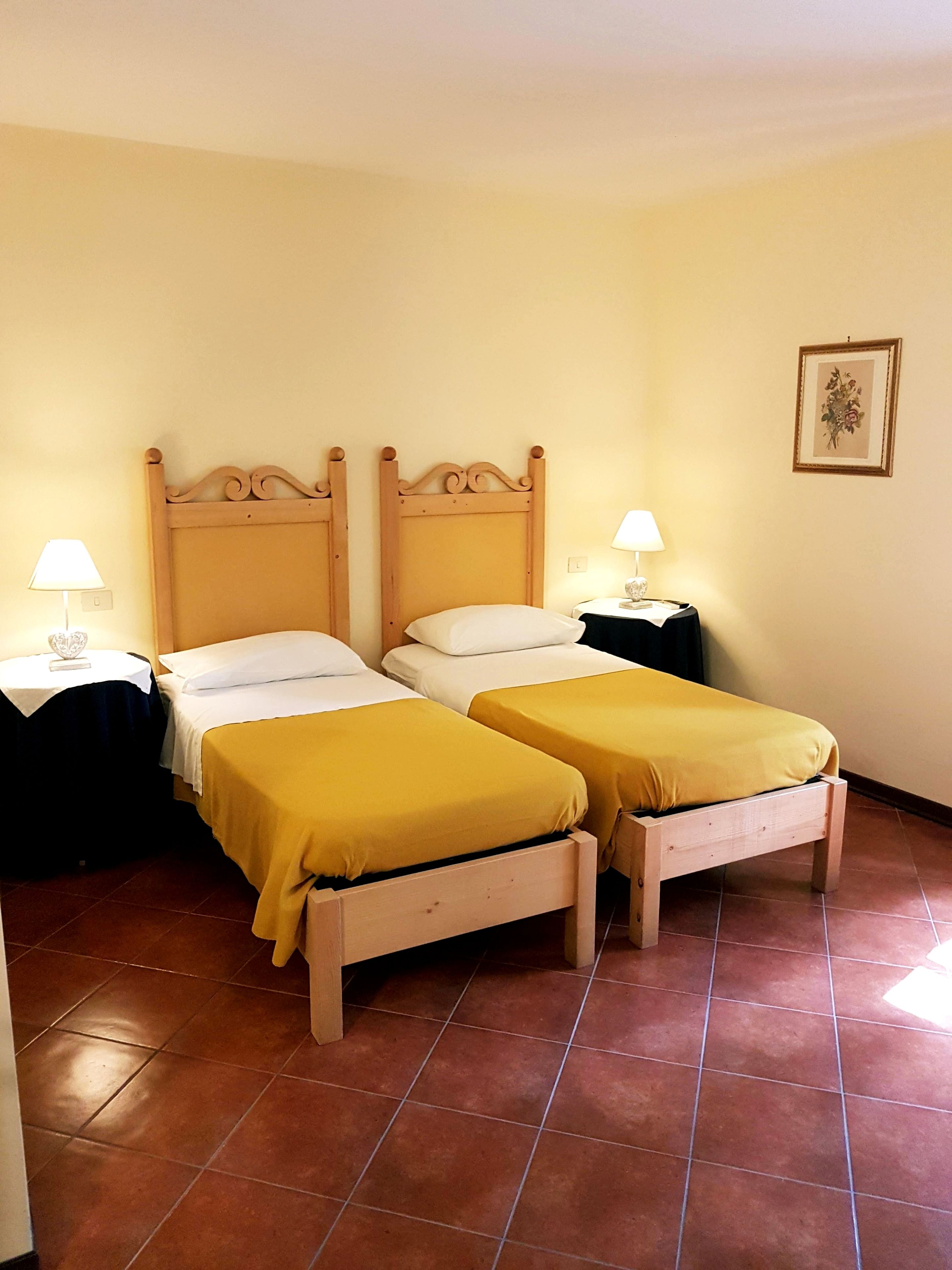 Een hotelkamer met twee enkele bedden, elk voorzien van een houten hoofdbord, witte beddengoed en een gele sprei. Er staan twee nachtkastjes met lampen en aan de muur hangt een ingelijst schilderij.
