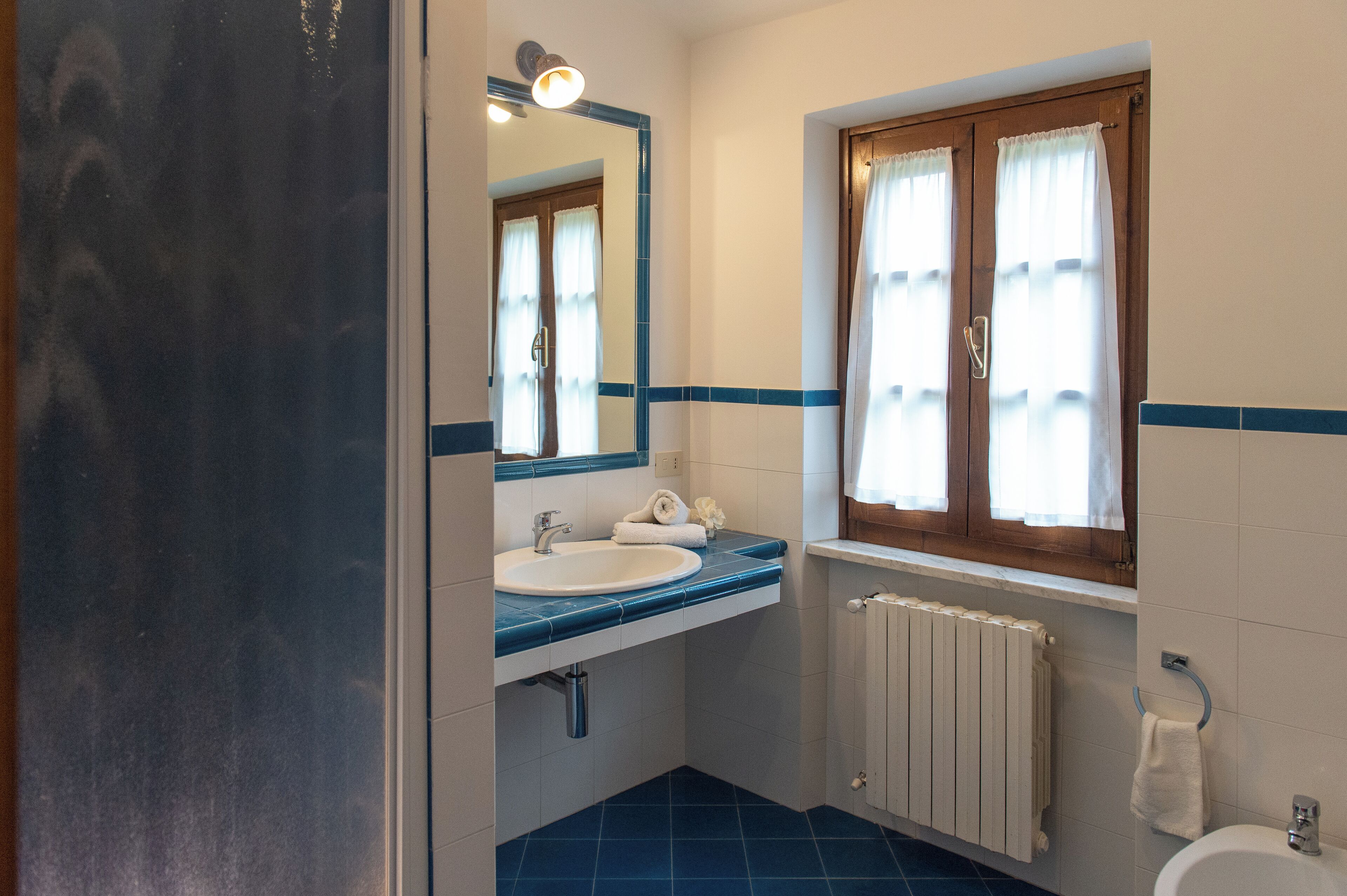 Un baño con lavamanos, espejo, ventana con cortinas y radiador.