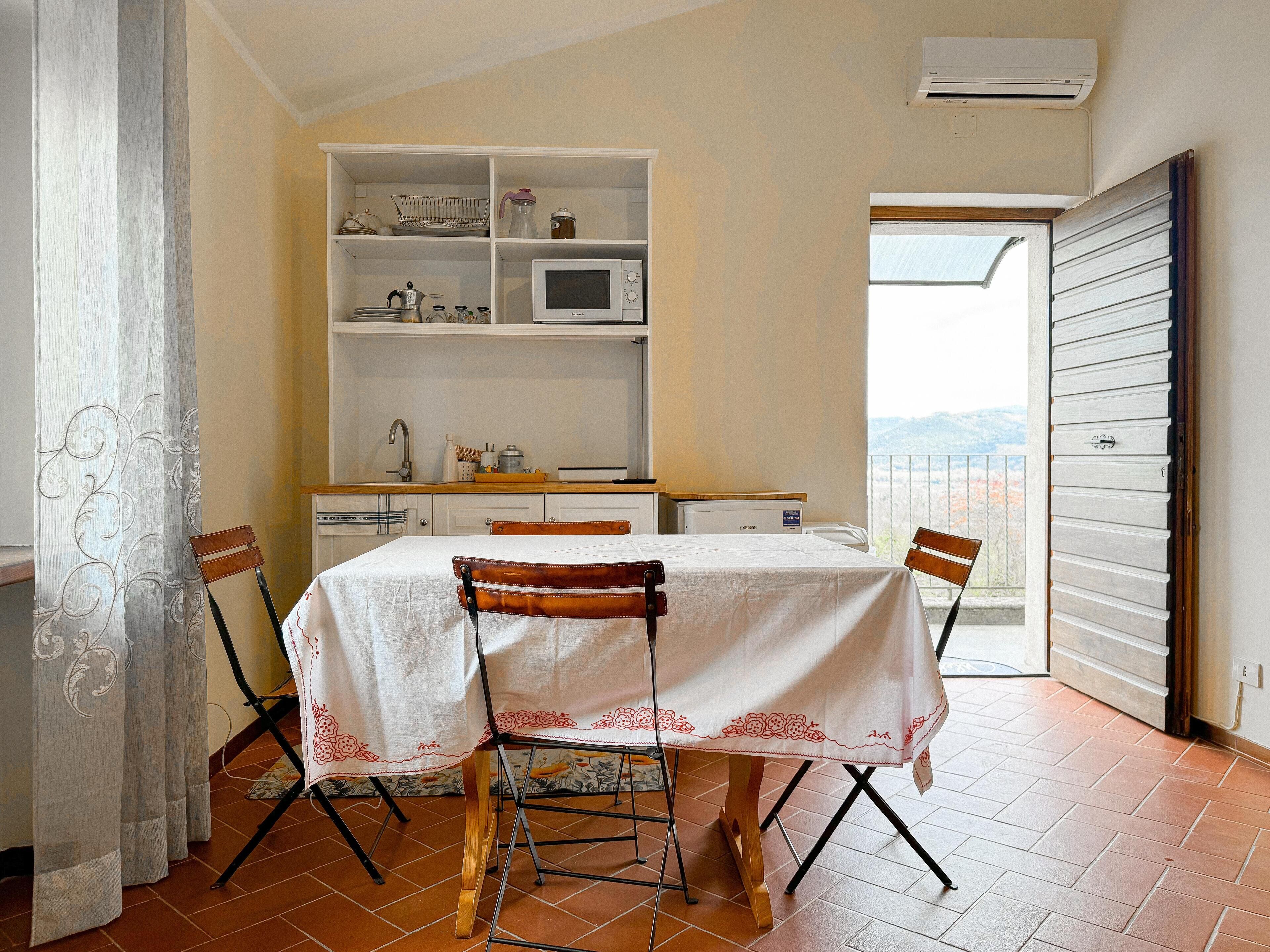 Comedor con una mesa dispuesta para cuatro, cocina al fondo y vistas al exterior a través de un ventanal abierto.