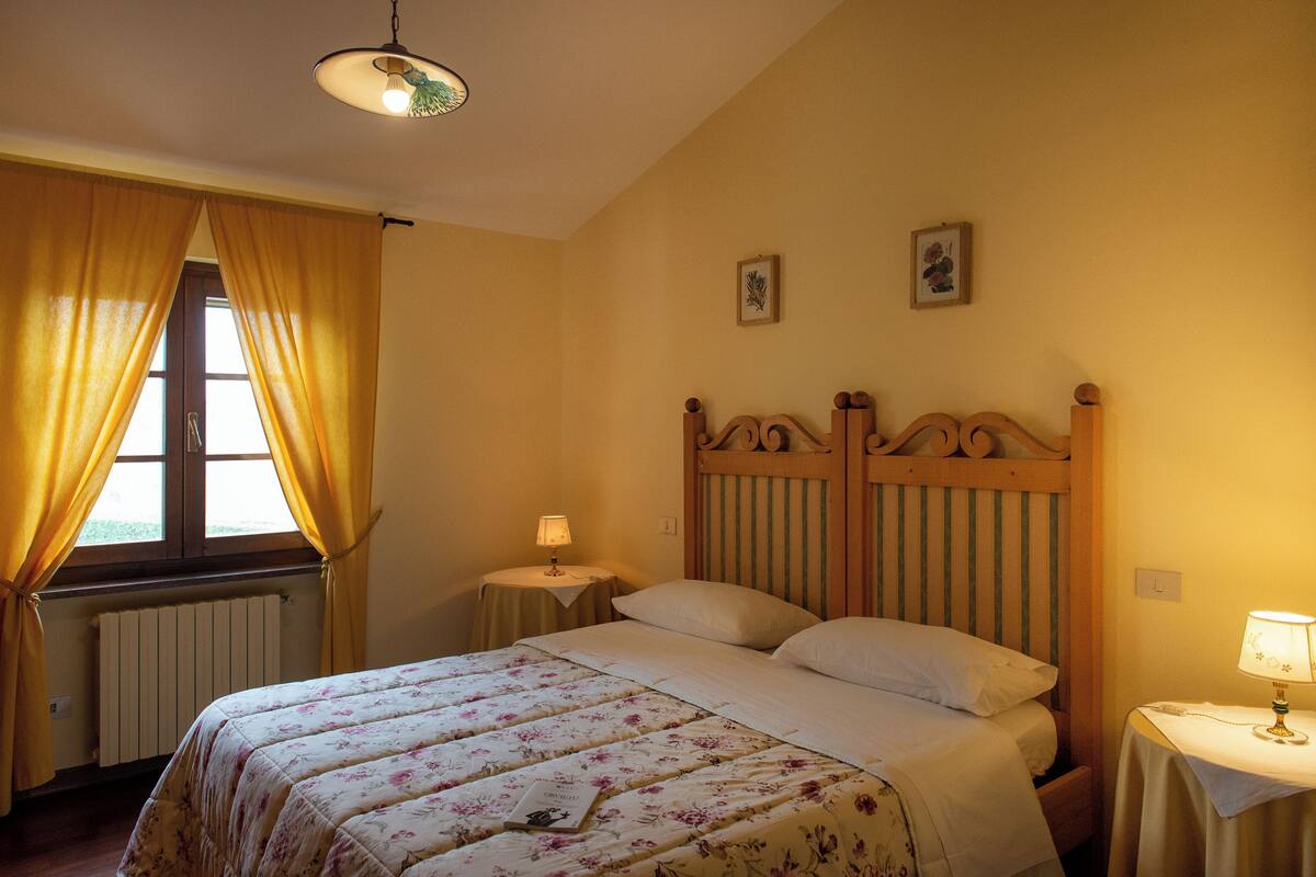 Quarto com cama, duas mesas de cabeceira, janela com cortinas, radiador e luz no teto.