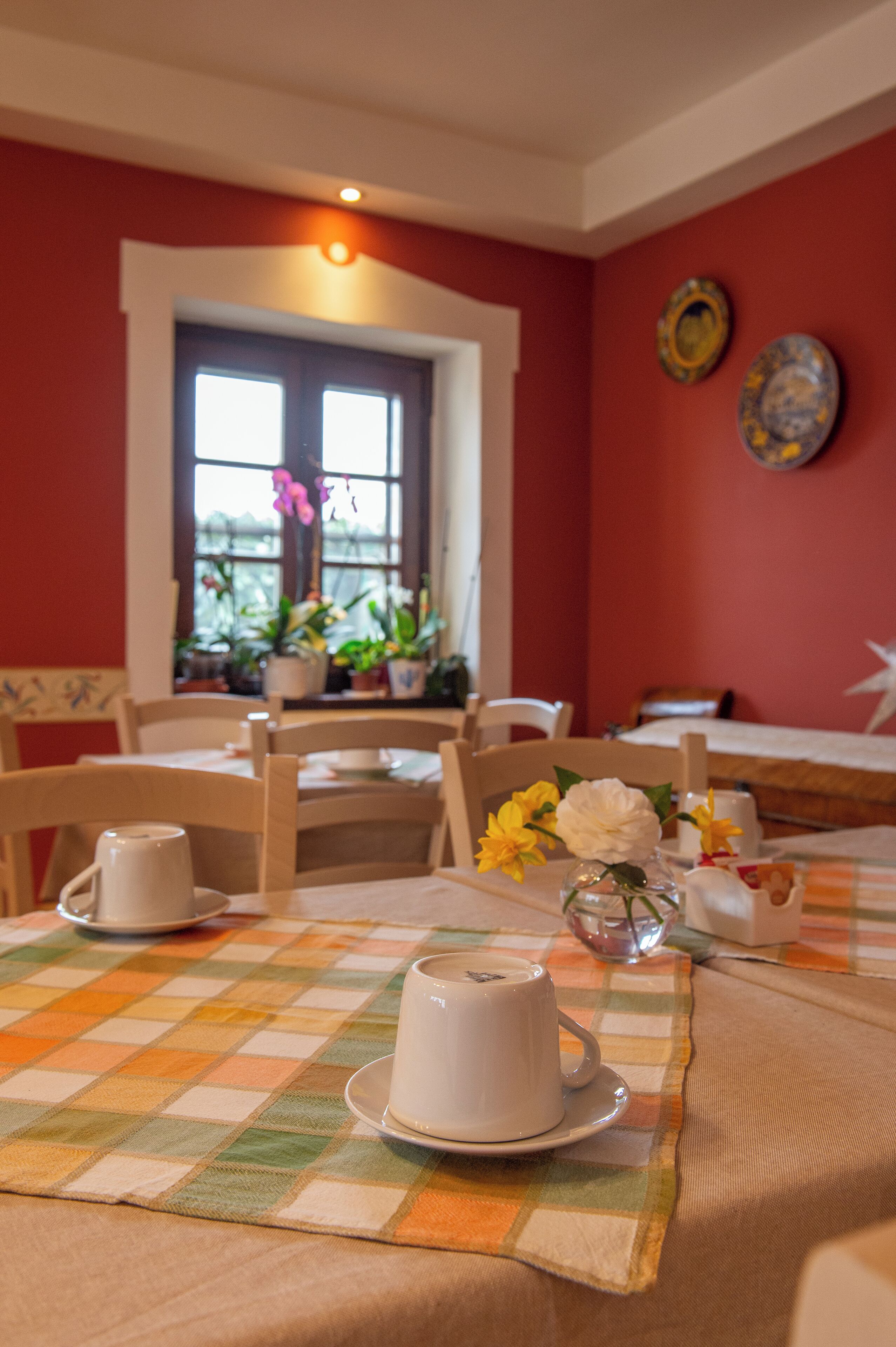 Un comedor con una mesa dispuesta para dos personas, con mantel a cuadros, una taza blanca y un jarrón con flores.