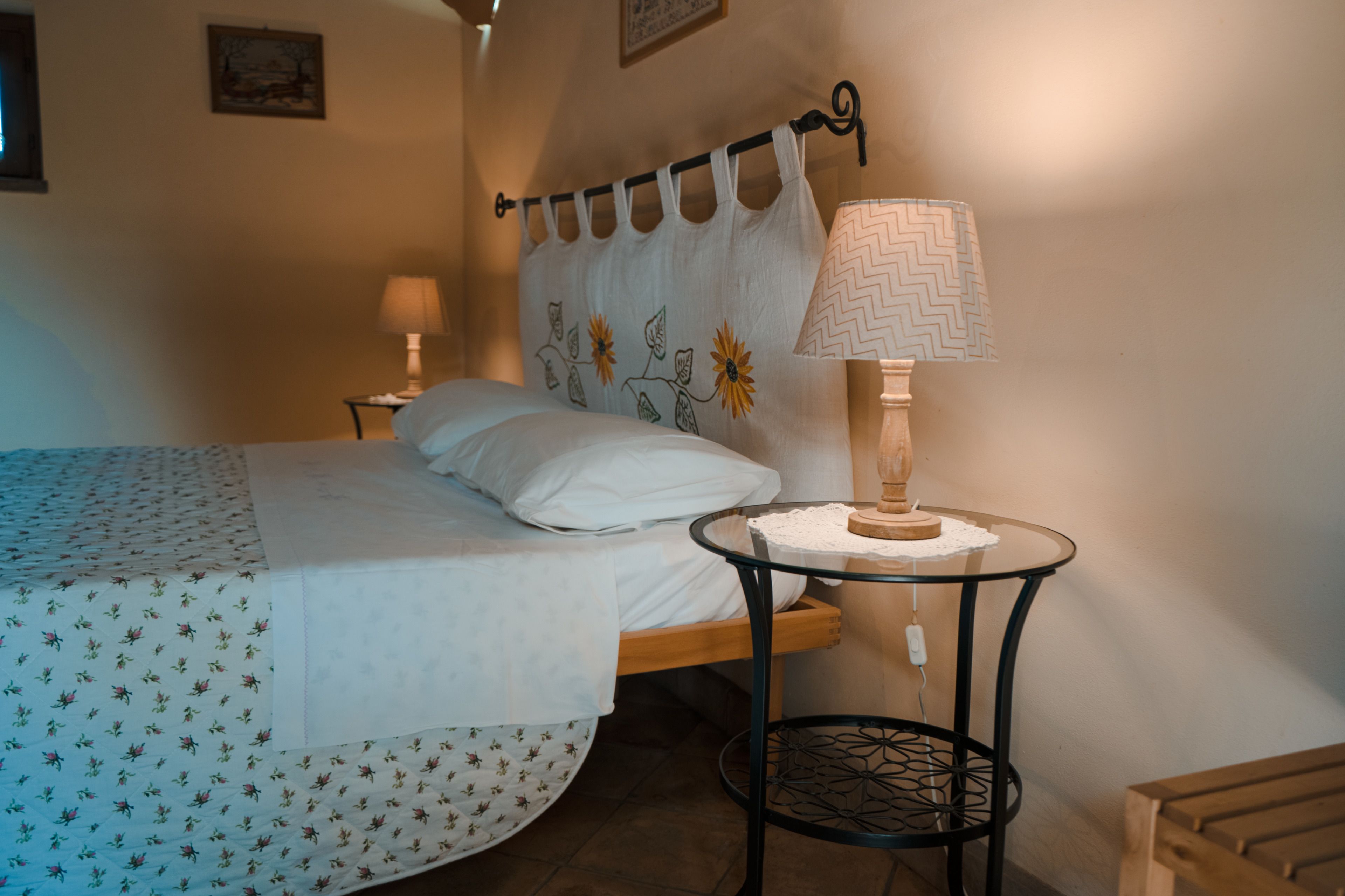 A neatly made bed with a floral bedspread, a bedside table with a lamp, and a wall-mounted curtain.