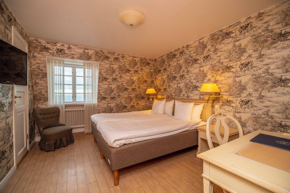A bedroom with a bed, two chairs, a desk, a window with curtains, and a wall with a floral pattern.