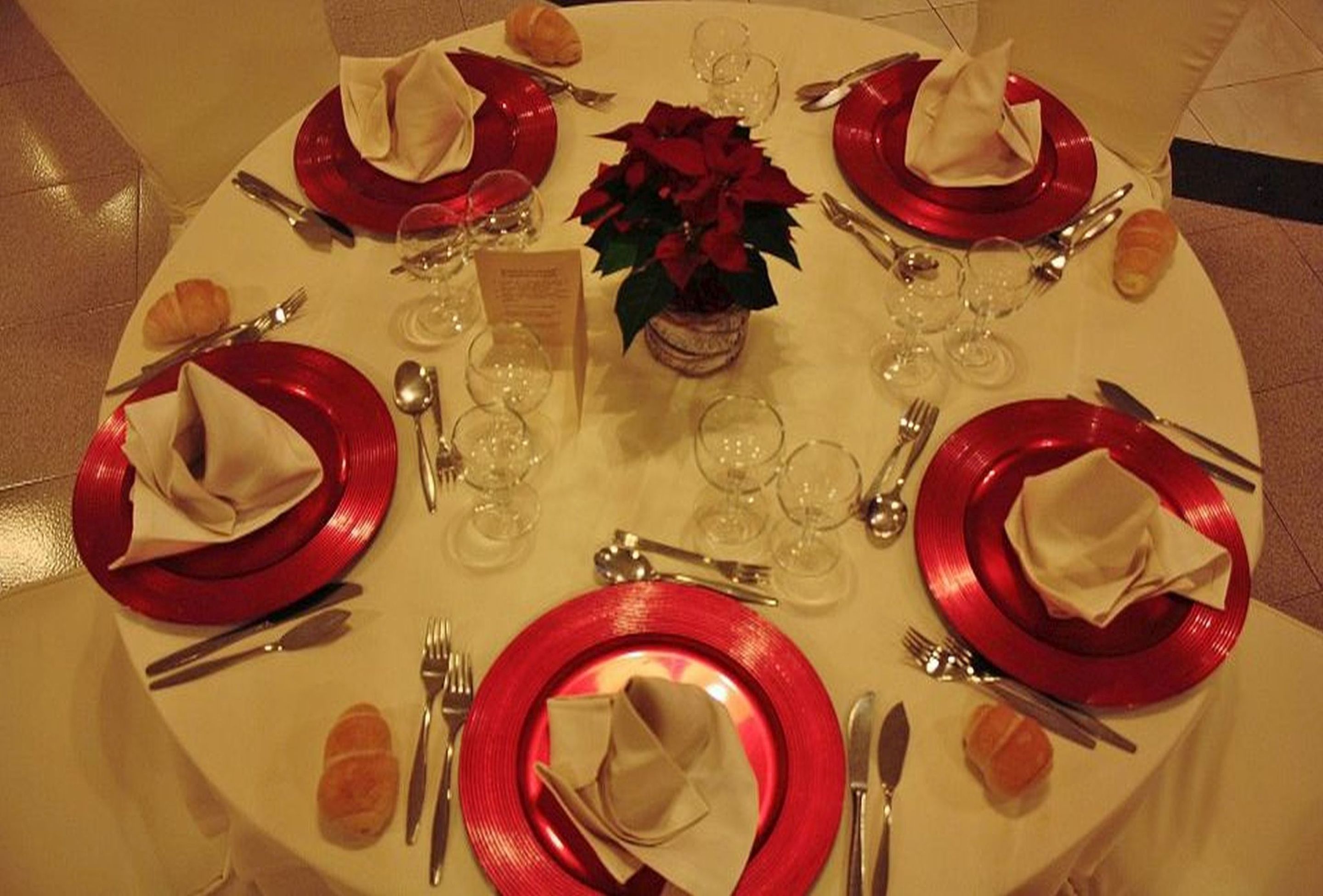 Une table de salle à manger ronde, dressée pour quatre personnes, avec des assiettes rouges, des serviettes blanches et un arrangement floral au centre.