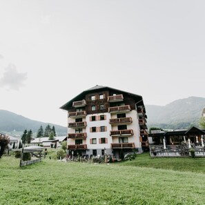 Ein mehrstöckiges Hotel mit Balkonen, gelegen in einer gebirgigen Gegend mit einer grünen Rasenfläche im Vordergrund.