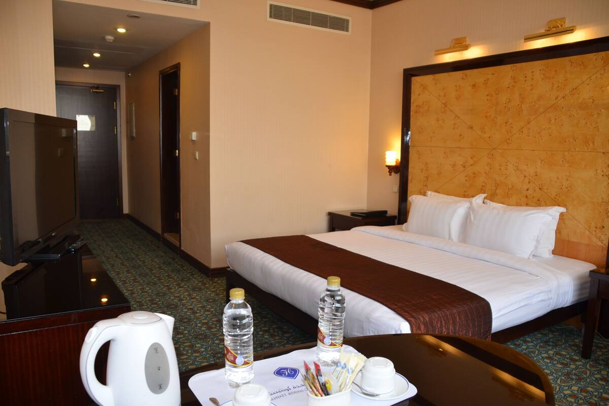 A hotel room with a bed, bedside table, and a desk with a kettle and water bottles.