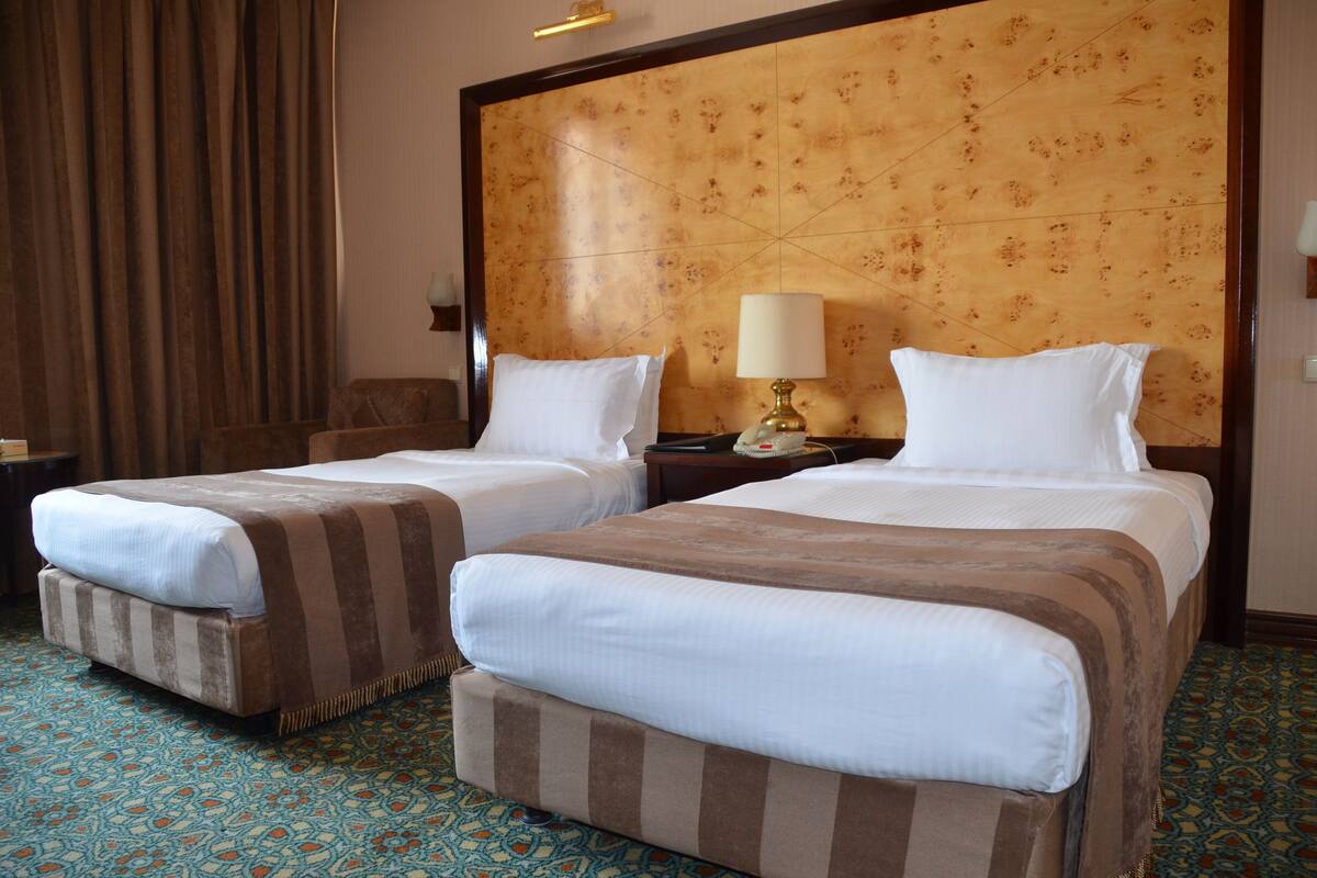 A hotel room with two beds, a wooden headboard, a patterned carpet, and a nightstand with a lamp.