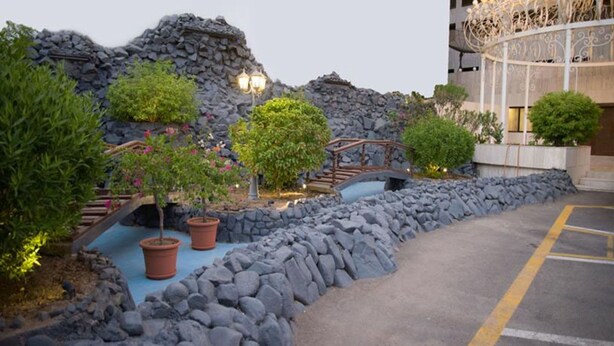 A landscaped garden with a stone wall, potted plants, a small bridge, and a parking area. - Al Hayatt Jeddah Hotel (Jeddah)
