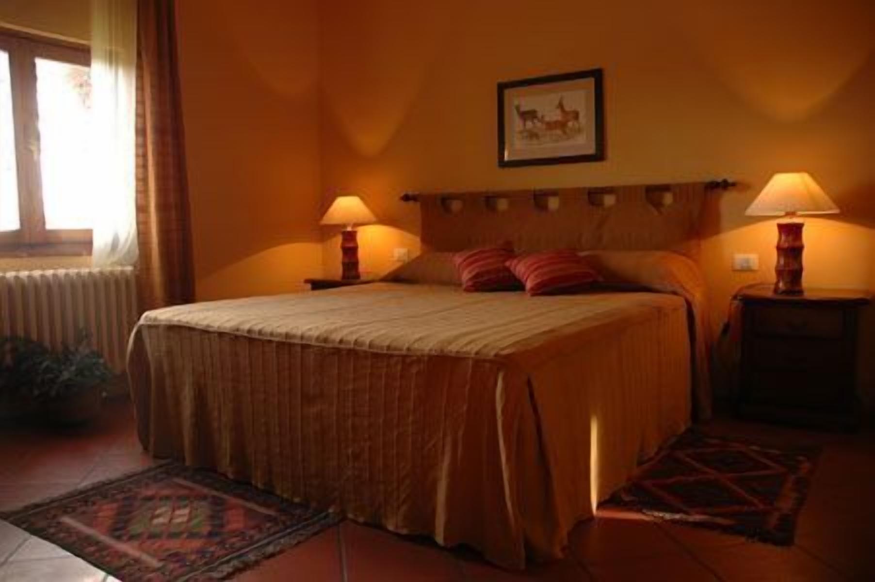 A neatly made bed with a headboard, two bedside lamps, a framed picture on the wall, and a window with curtains.