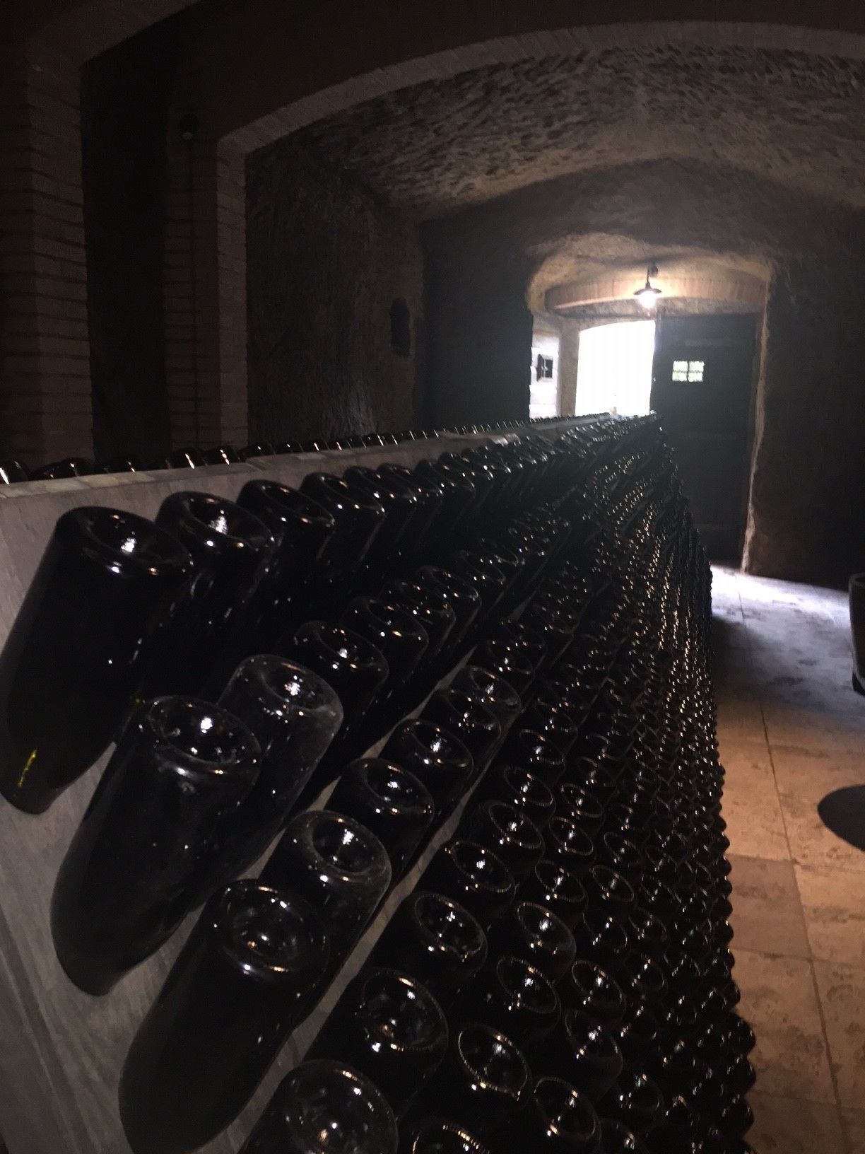 Una hilera larga de copas de vino vacías alineadas en un pasillo tenuemente iluminado, con paredes de ladrillo y una puerta al final.