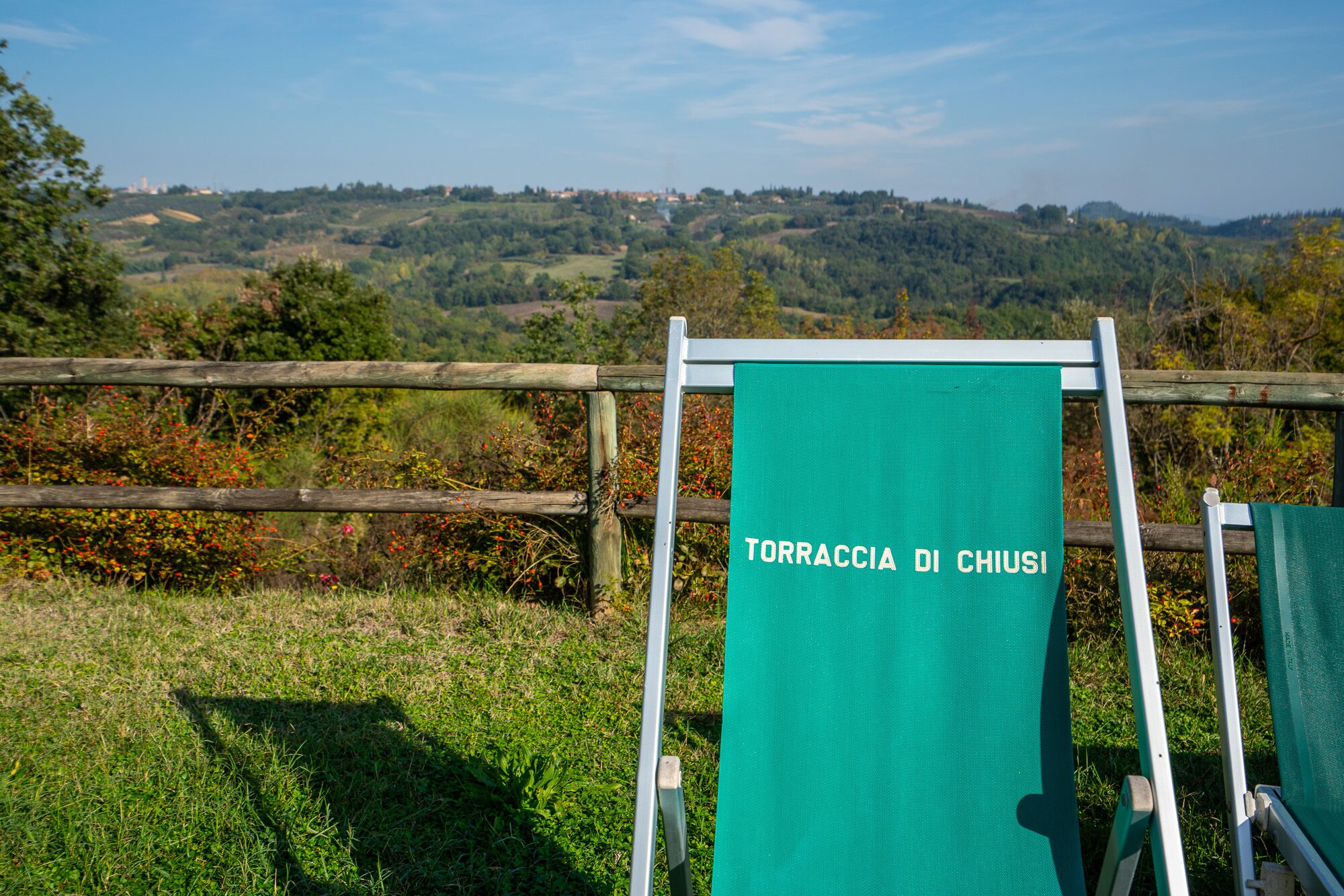 A green sign with the text "TORRACCI DI CHIUSI" on a metal frame, set against a rural landscape with hills and trees.