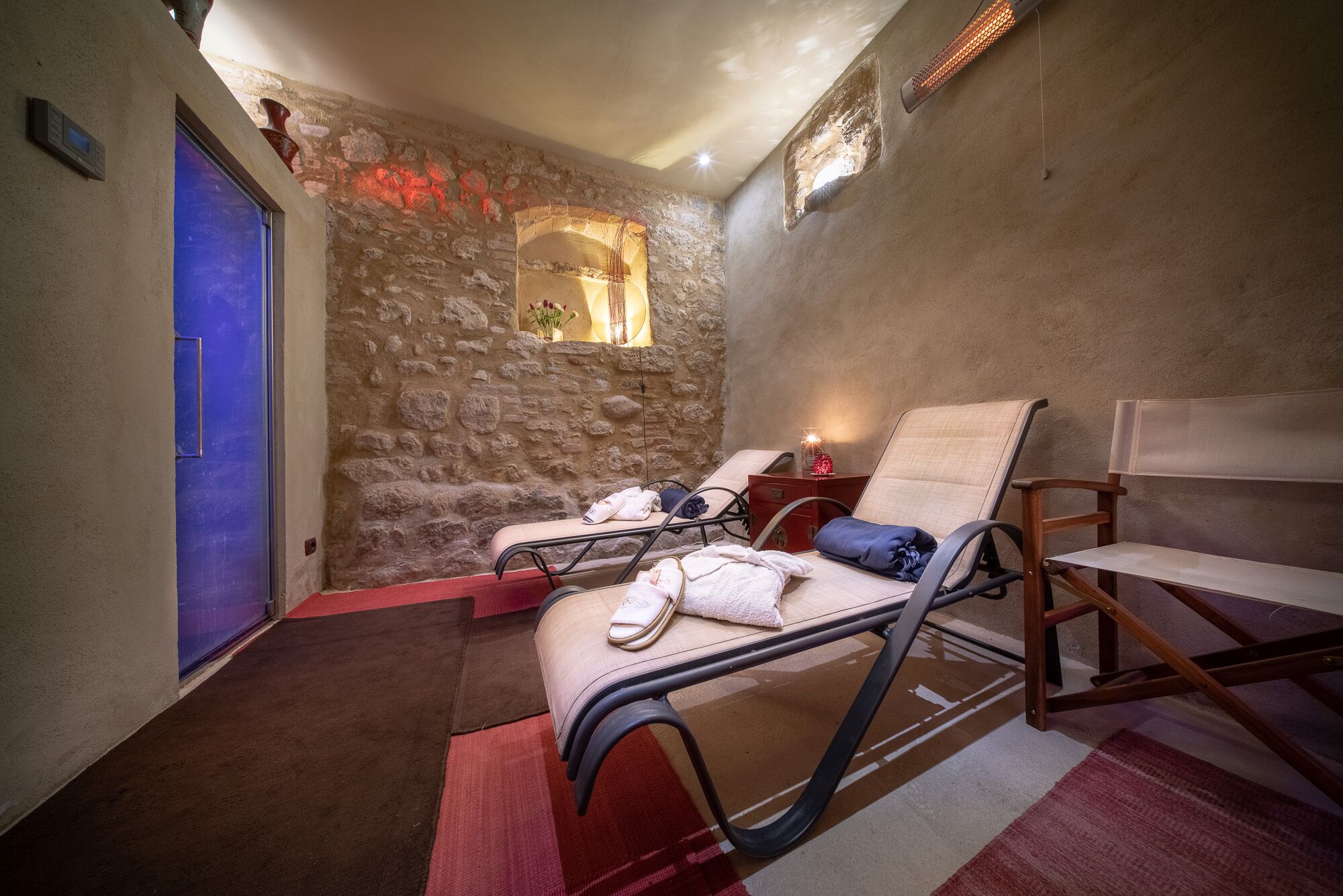 Une salle de spa équipée de deux tables de massage, d’un mur en pierre, d’une porte bleue et d’une petite fenêtre éclairée de lumière rouge.