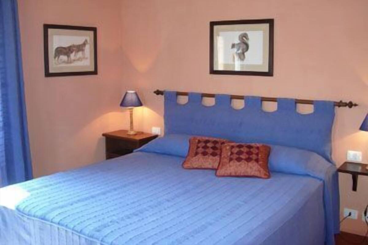 A bedroom with a bed, two framed pictures on the wall, a nightstand with a lamp, and a blue headboard.