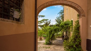 Un arco de entrada da a un patio con suelo de piedra, plantas en macetas y un edificio con techo de tejas.