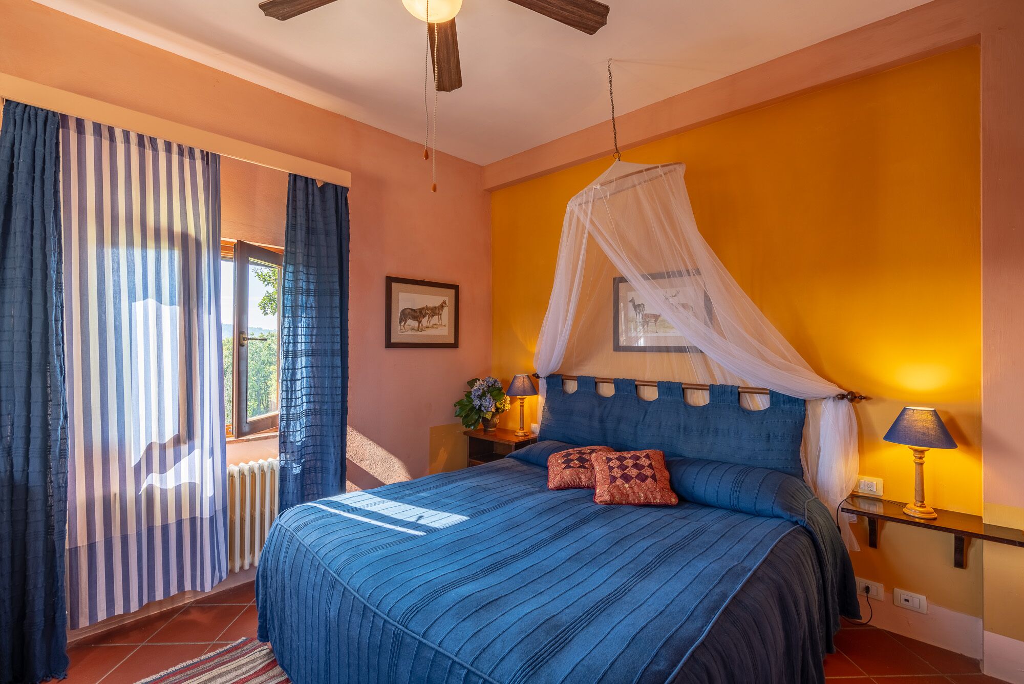 A bedroom with a bed, a canopy, a nightstand with a lamp, a picture on the wall, and a window with curtains.