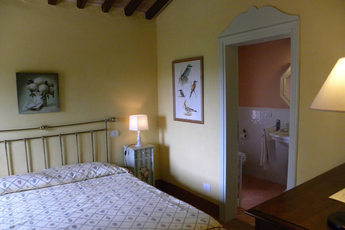 A bedroom with a bed, a bedside table with a lamp, a framed picture on the wall, and a bathroom visible through an open door.