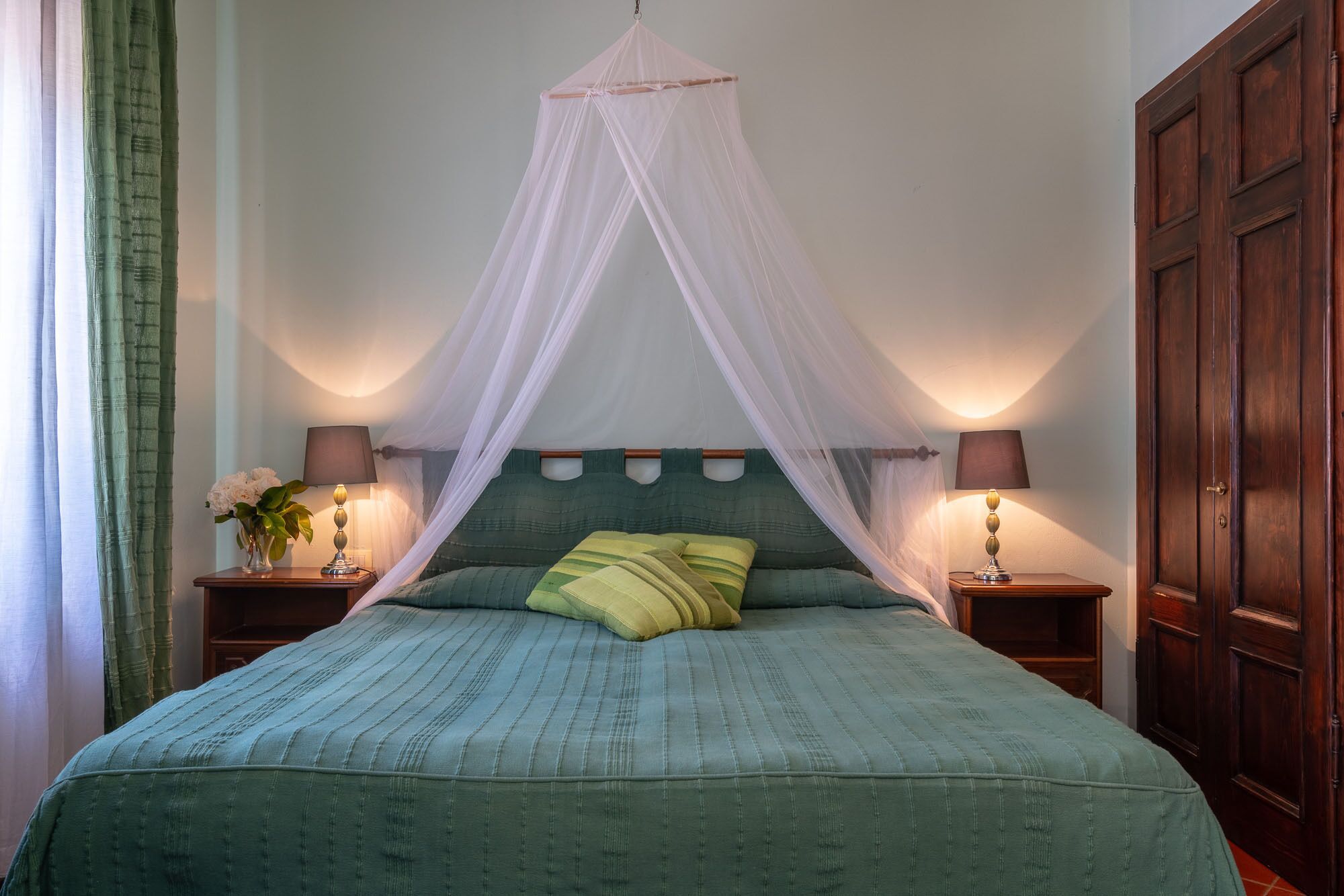 A bedroom with a canopy bed, two bedside tables with lamps, a wooden wardrobe, and a green-striped bedspread.