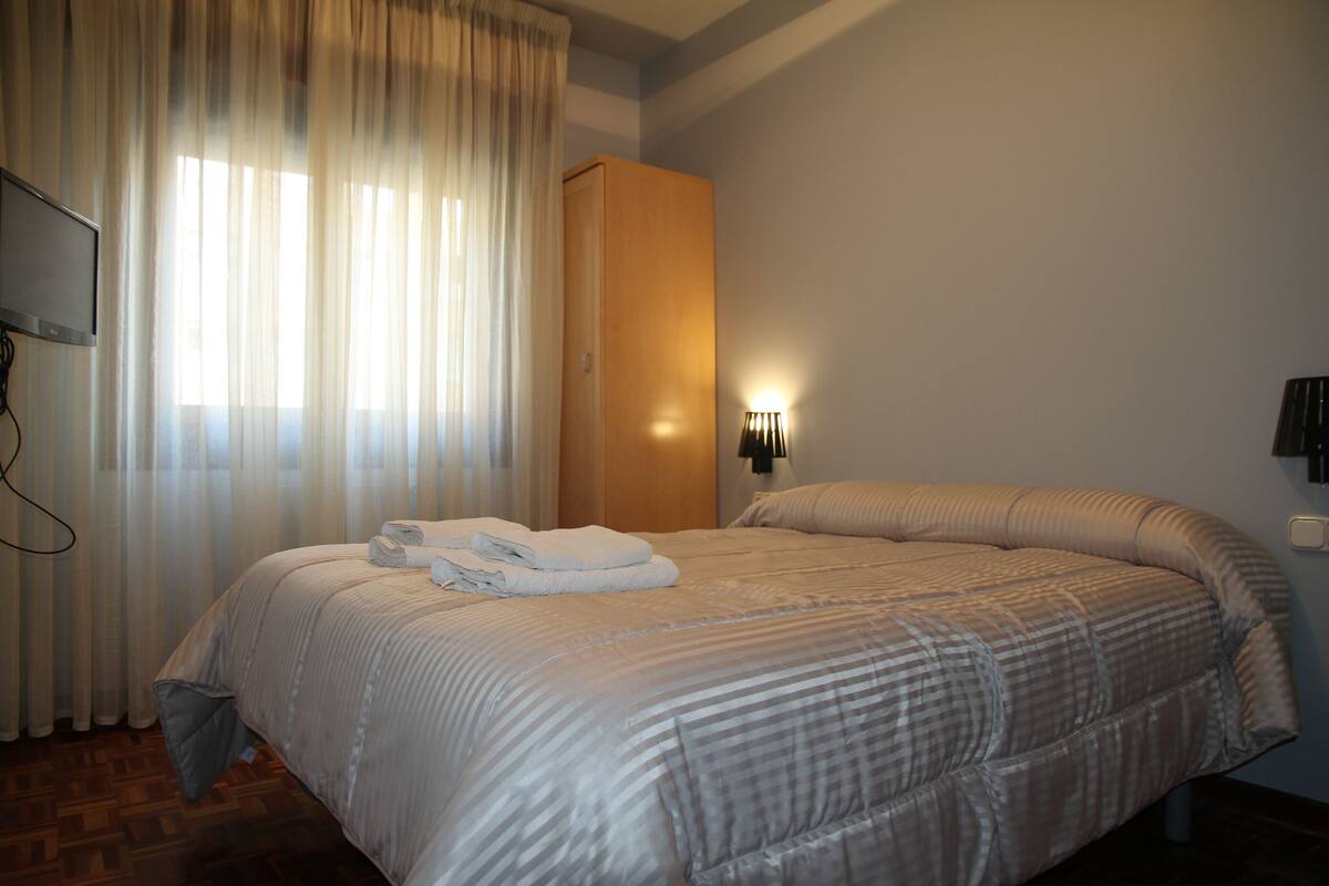 A neatly made bed with a striped blanket, a wooden wardrobe, a wall-mounted TV, and a bedside lamp.
