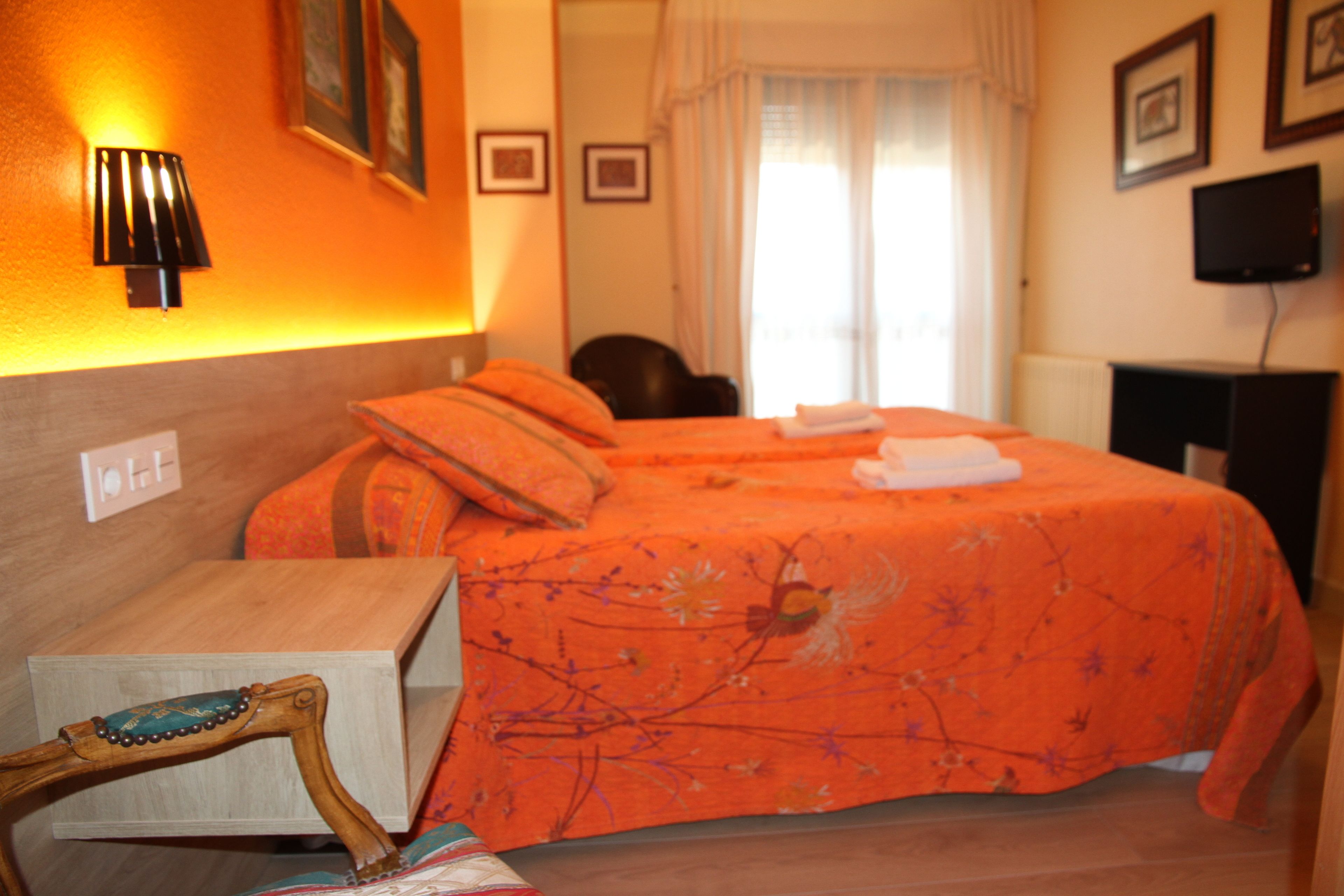 Habitación de hotel con cama, mesita de noche de madera, lámpara, televisor y ventana con cortinas.