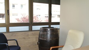 A room with a wooden floor, a large wooden barrel, a black chair, a white chair, and a window with a view of buildings and trees. - Hostal Acella (Pamplona)