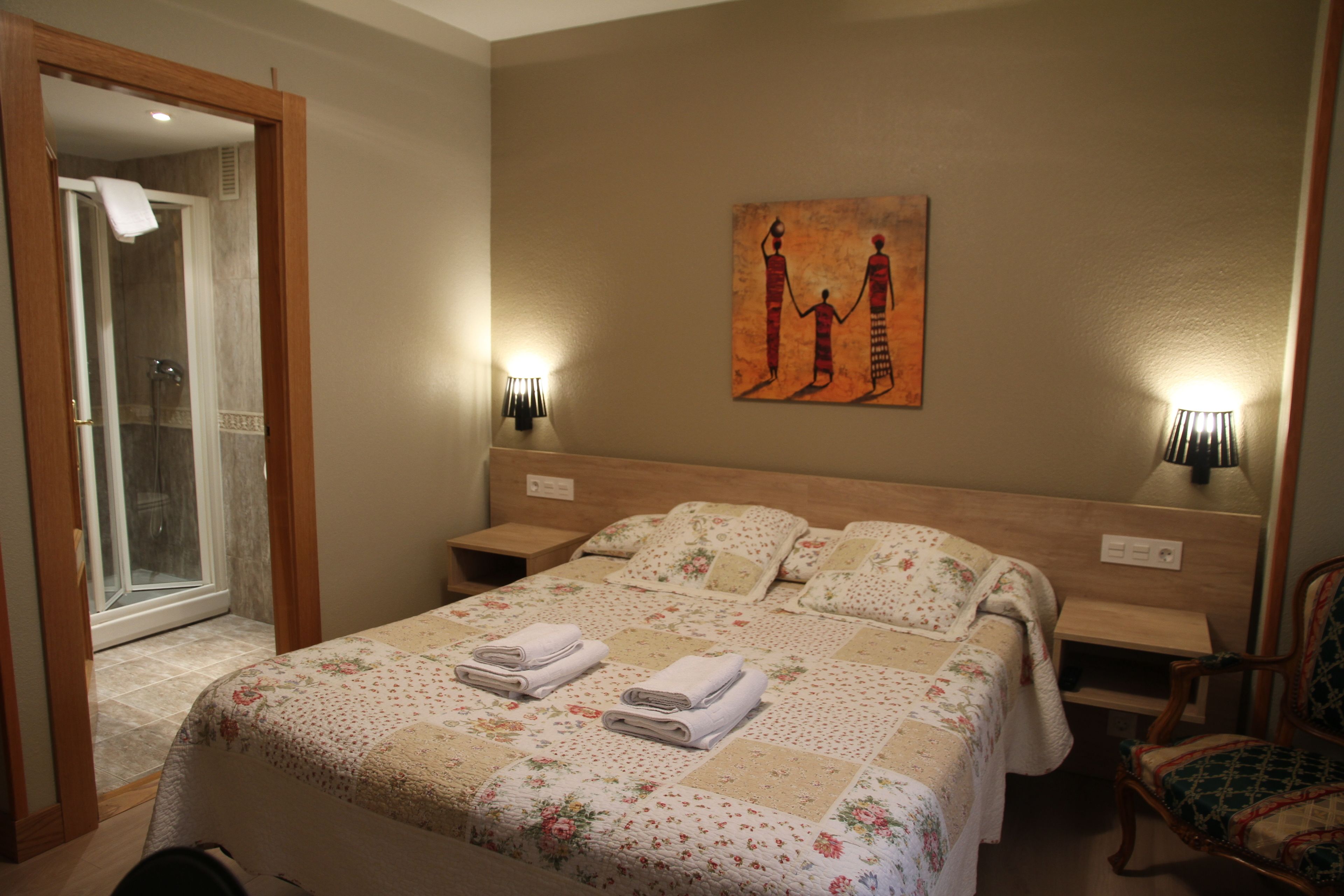Habitación de hotel con una cama grande, mampara de vidrio en la ducha, un cuadro en la pared y dos lámparas de noche.