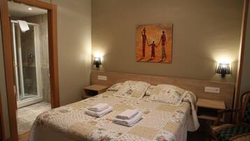 Habitación de hotel con una cama grande, mampara de vidrio en la ducha, un cuadro en la pared y dos lámparas de noche.