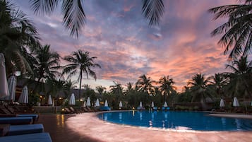 Una piscina con sillas de descanso y sombrillas, rodeada de palmeras, bajo un cielo al atardecer.