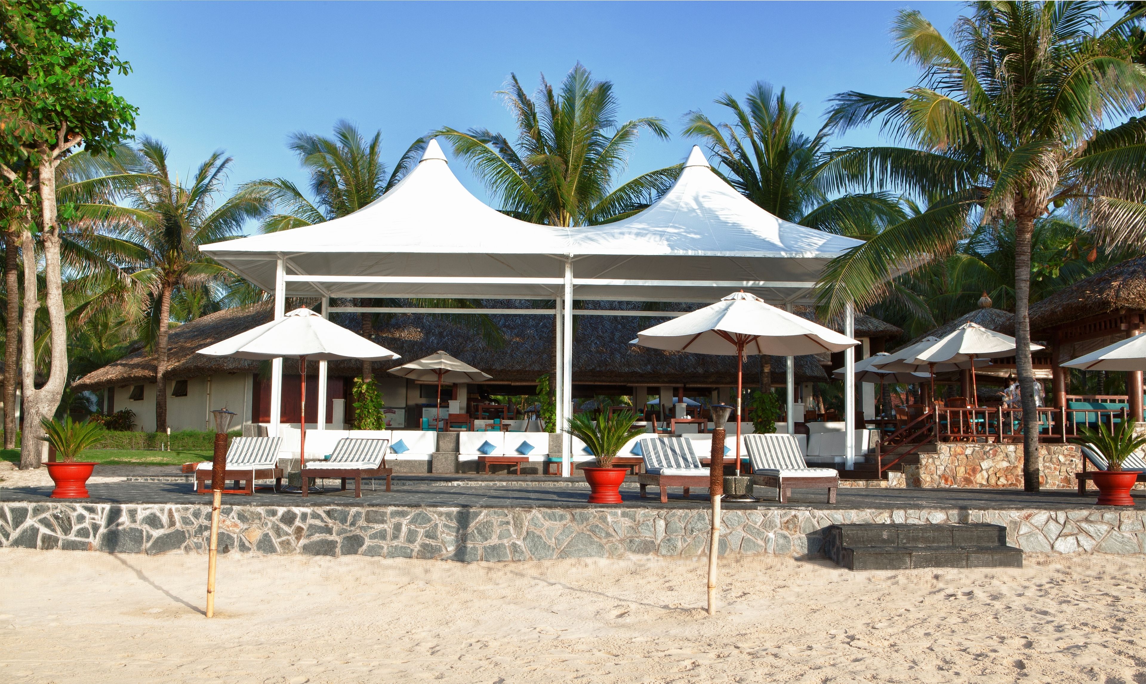 Un resort frente a la playa con una zona de estar techada, palmeras y un muro de piedra.