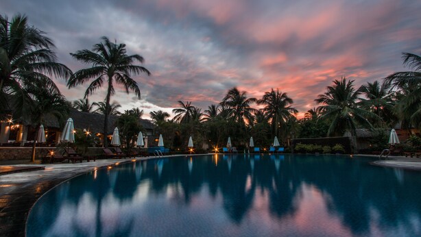 A pool area with lounge chairs and umbrellas, surrounded by palm trees, under a twilight sky. - Blue Ocean Resort (Mui Ne)