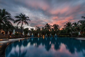A pool area with lounge chairs and umbrellas, surrounded by palm trees, under a twilight sky.