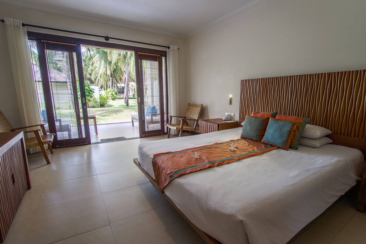 A hotel room with a large bed, a small table, a chair, and a sliding glass door leading to an outdoor area.