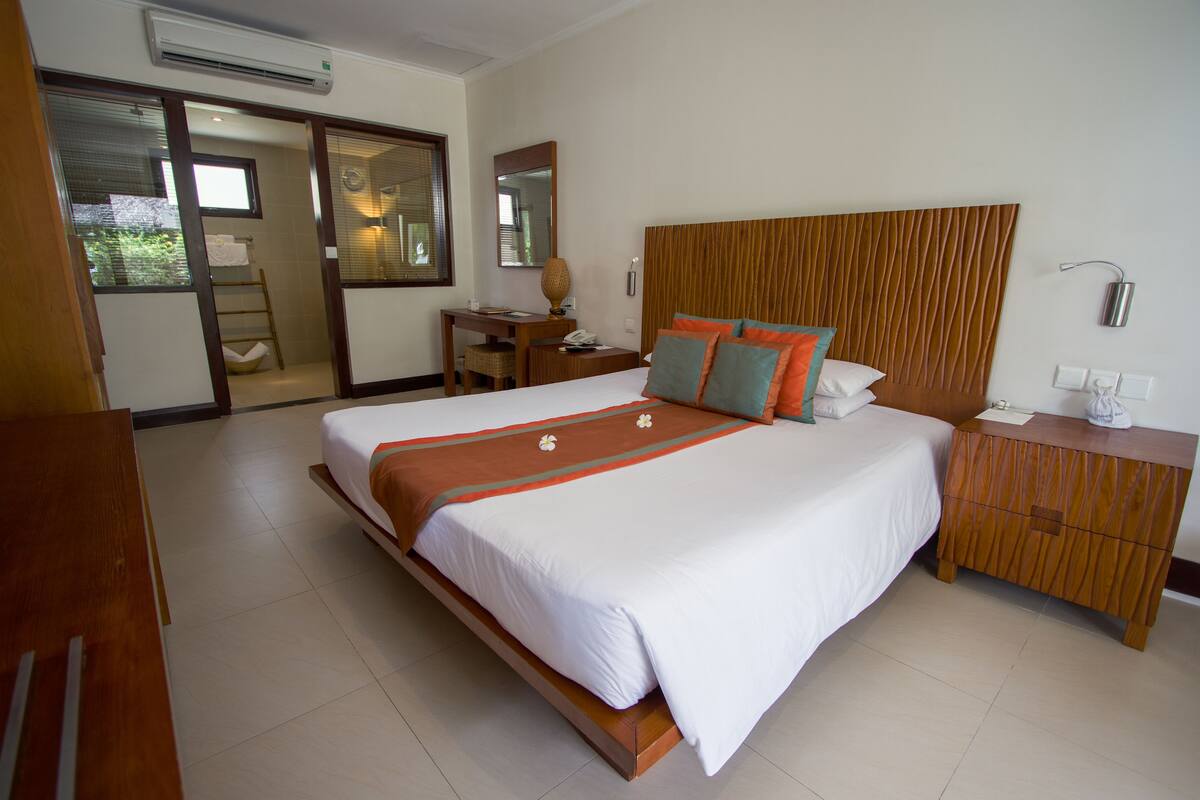 A hotel room with a large bed, a wooden headboard, a desk, and a bathroom visible through a sliding door.